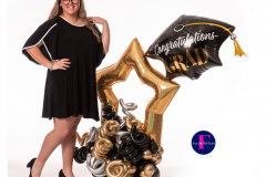 A woman in a black dress is standing next to a graduation cap balloon.