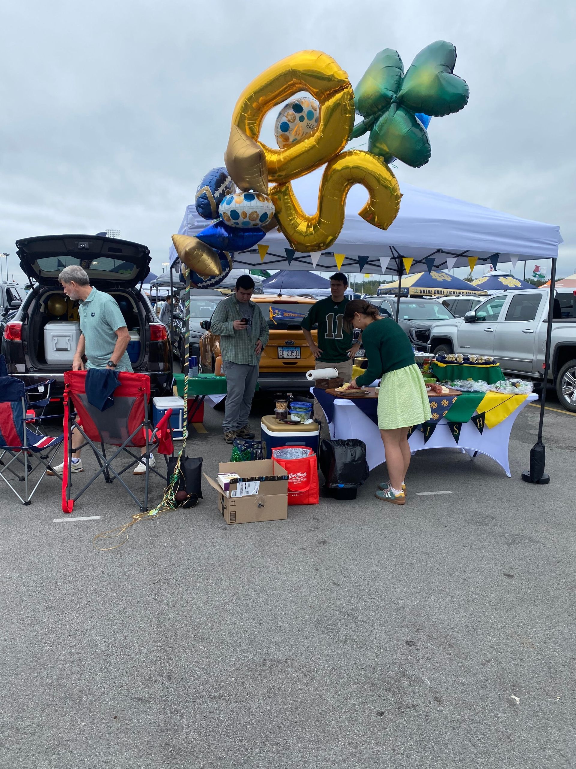 Tailgating scene with balloons; people near a table with food, drinks, and a tent in a parking lot.