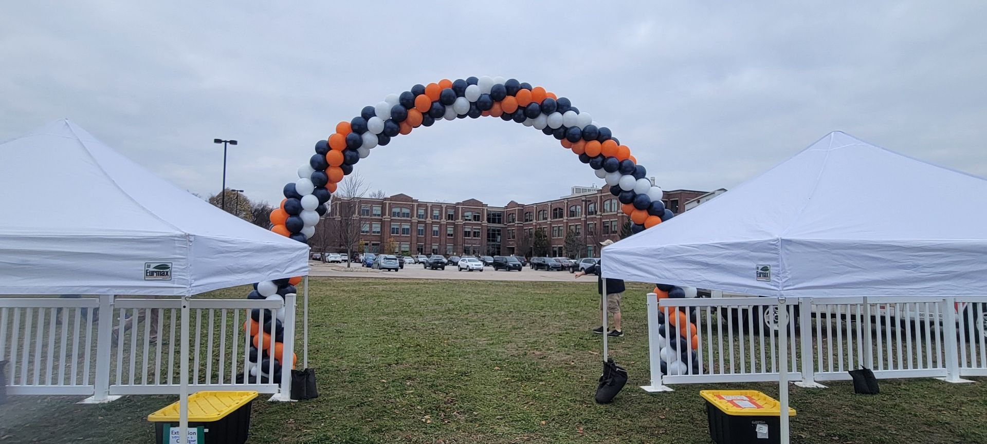Two white tents flank a balloon arch in a park setting. Balloons are orange, navy, and white.