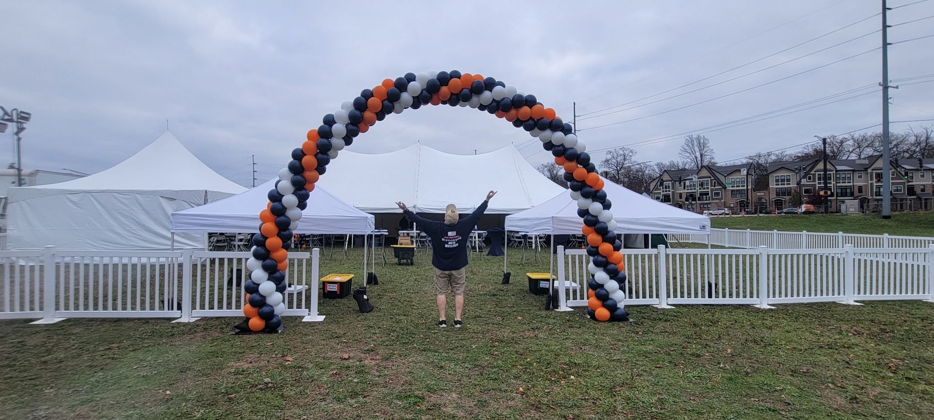Person stands under a balloon arch, arms outstretched. White tents and fence in the background, cloudy sky.