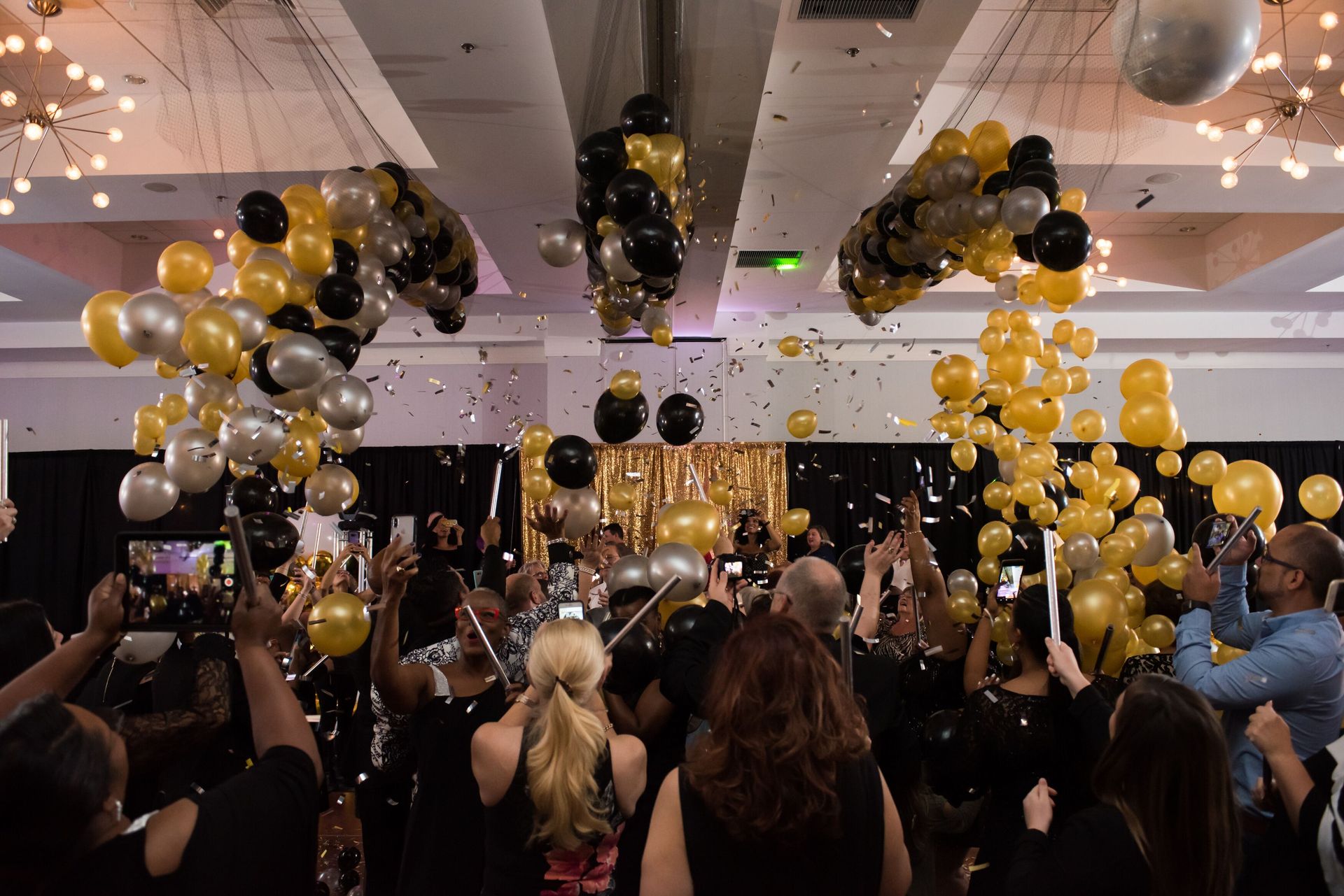 A crowd of people are standing in a room with balloons and confetti falling from the ceiling. Balloon Drop