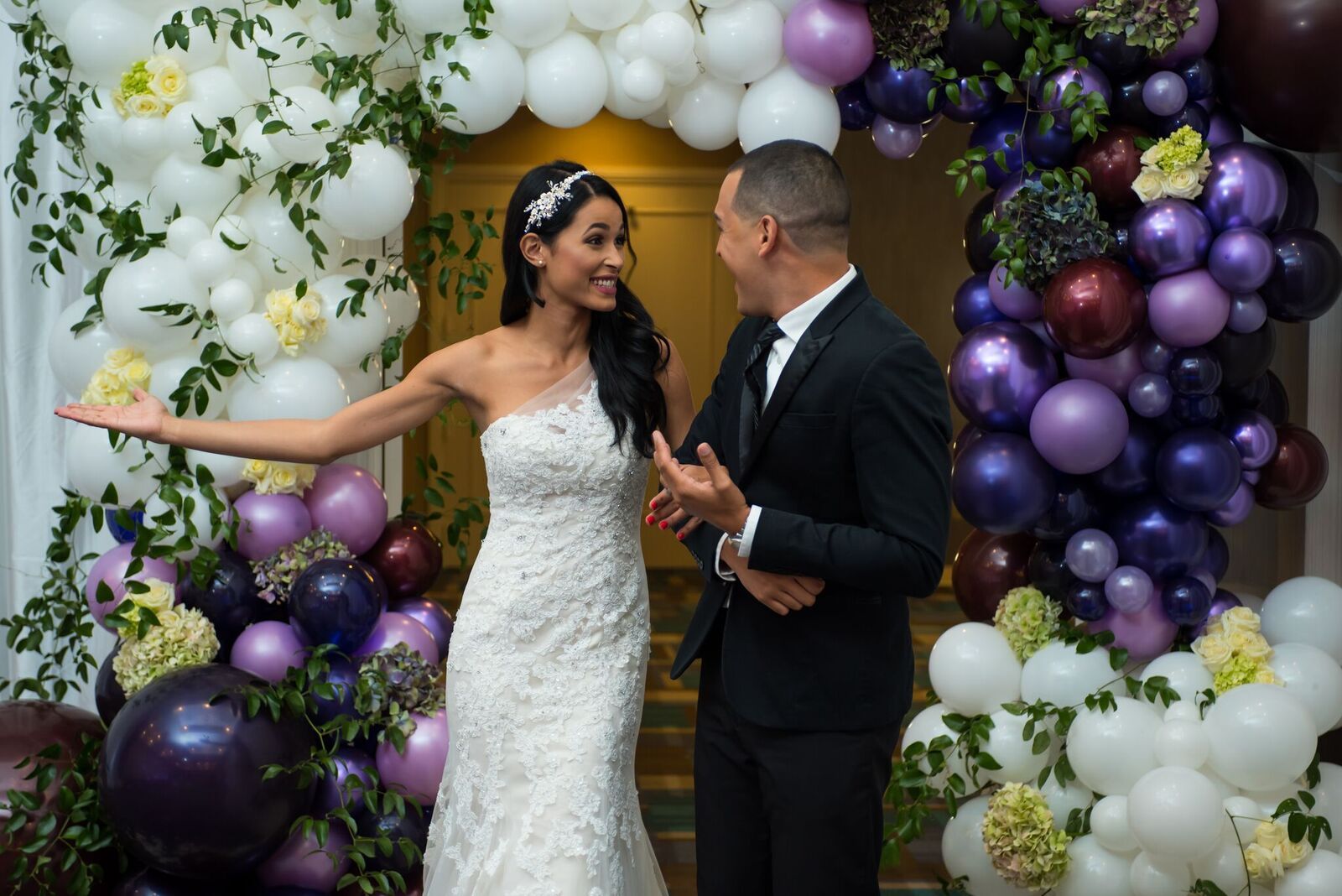 A bride and groom are standing in front of a wall of balloons.