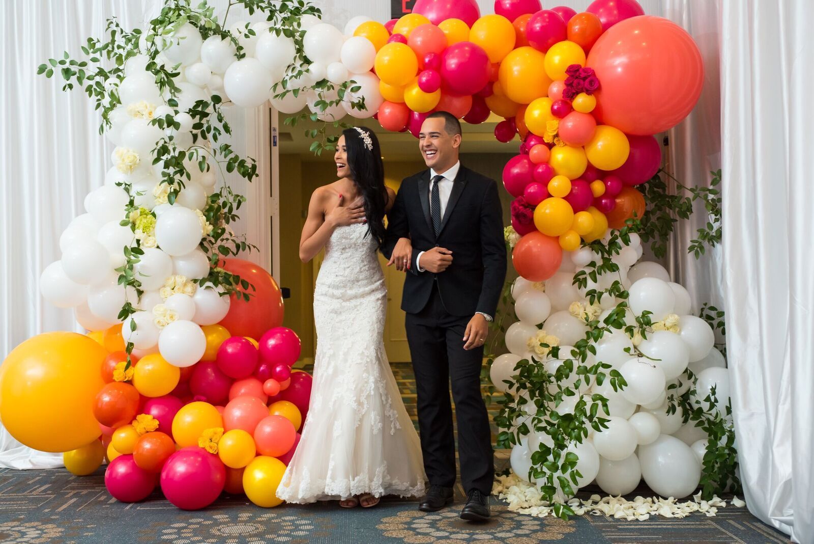 A bride and groom are walking under a balloon arch.