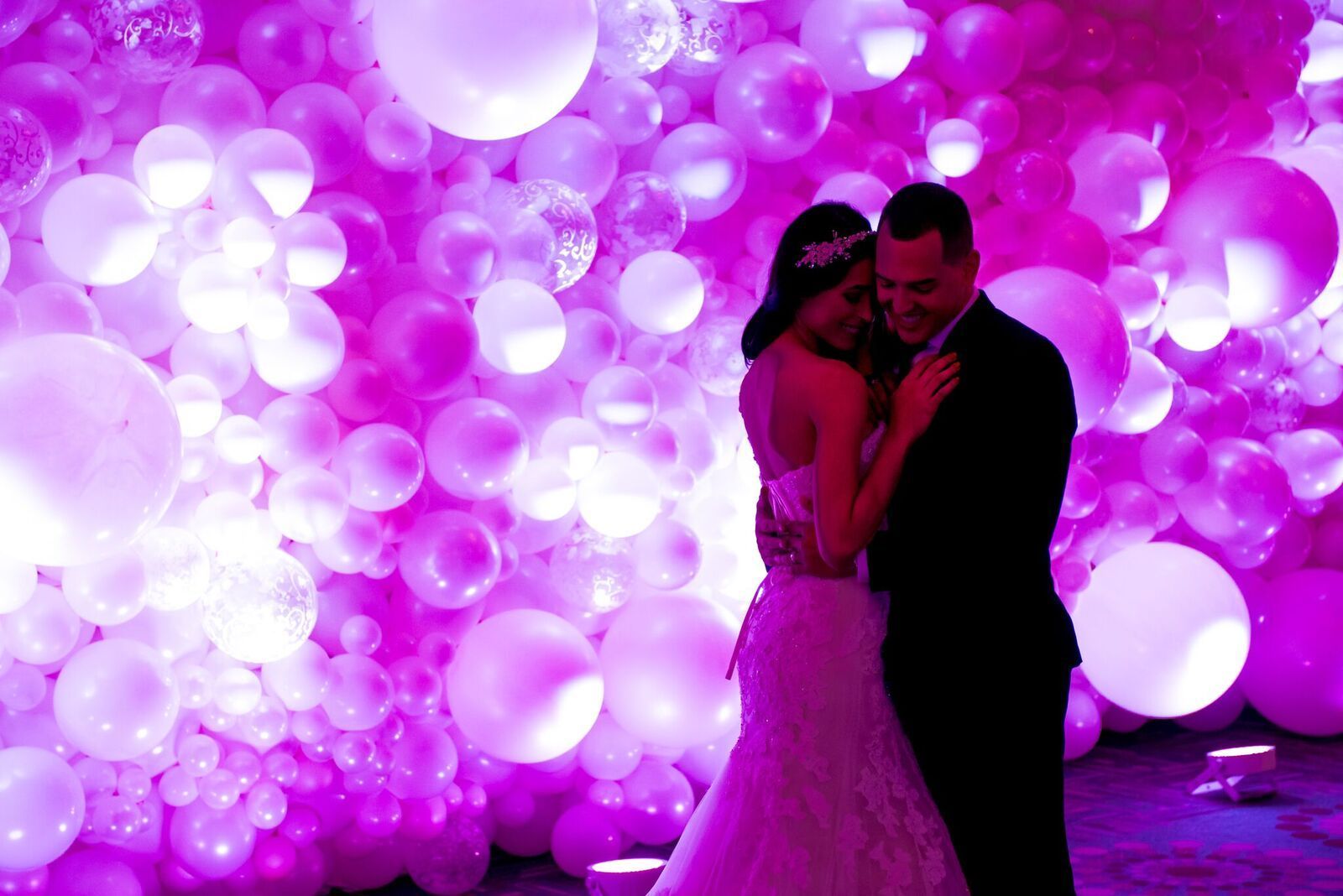 A bride and groom are standing in front of a wall of pink balloons.
