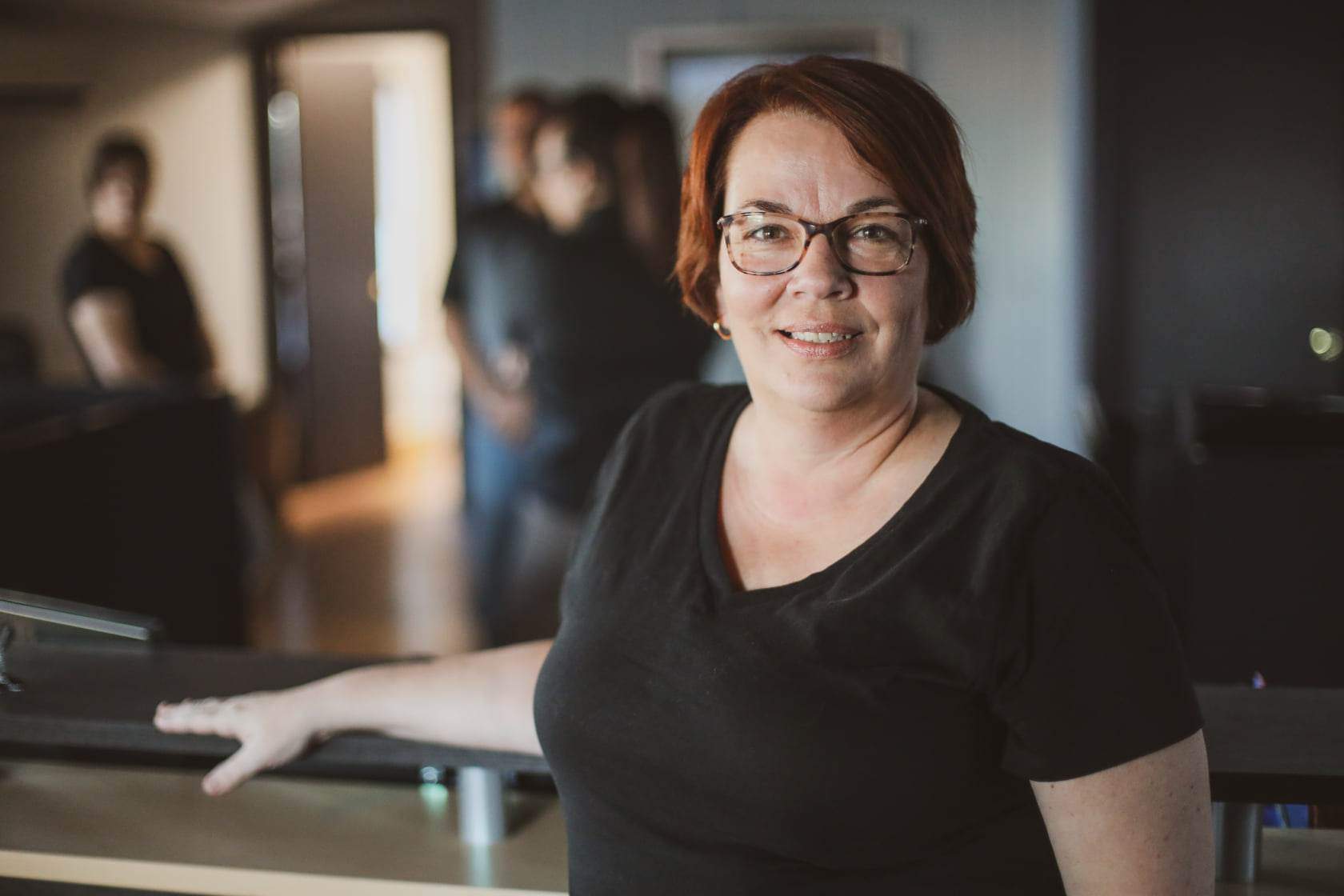 Woman with red hair and glasses in a black shirt smiles, leaning on a counter, with others blurred in the background.