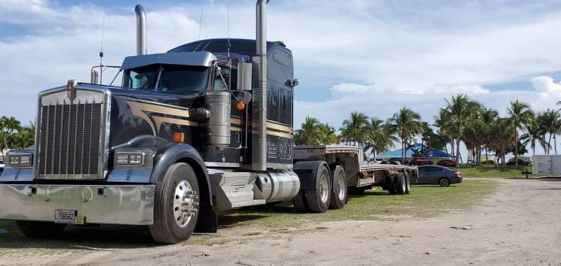 Un semi-remorque Kenworth noir est garé à l'extérieur sur une pelouse, avec une remorque à plateau attachée. Des palmiers et un ciel bleu en arrière-plan.