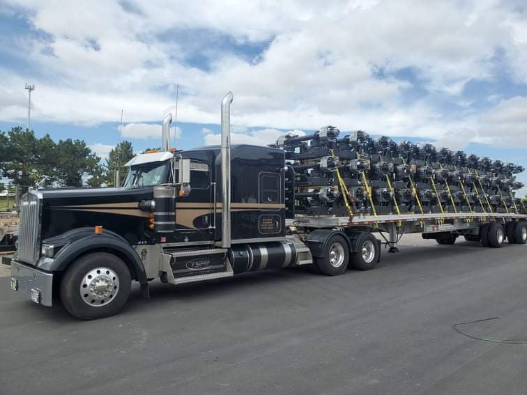 Un semi-remorque noir transporte des rangées d'équipements noirs sur une remorque à plateau sous un ciel nuageux.