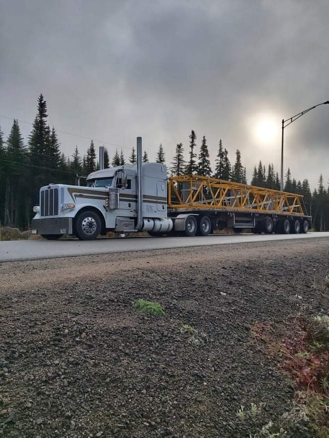 Un semi-remorque blanc avec une remorque plateau jaune transporte une longue structure jaune sur une route grise bordée d'arbres. Ciel couvert.