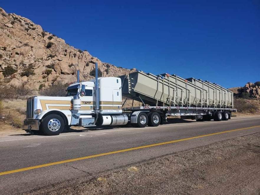 Un semi-remorque blanc et beige transportant plusieurs conteneurs gris circule sur une autoroute avec des montagnes rocheuses en arrière-plan, sous un ciel bleu.