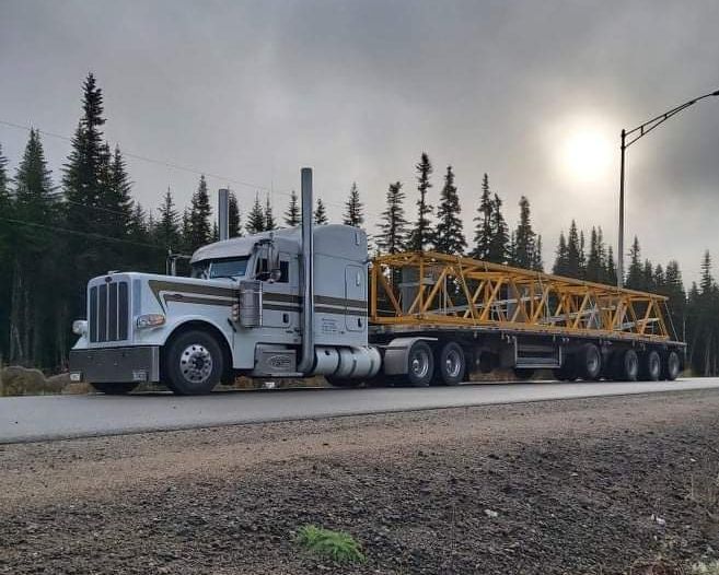 Semi-camion blanc avec une remorque à plateau jaune transportant un long cadre métallique sur une route sous un ciel nuageux, des arbres en arrière-plan.