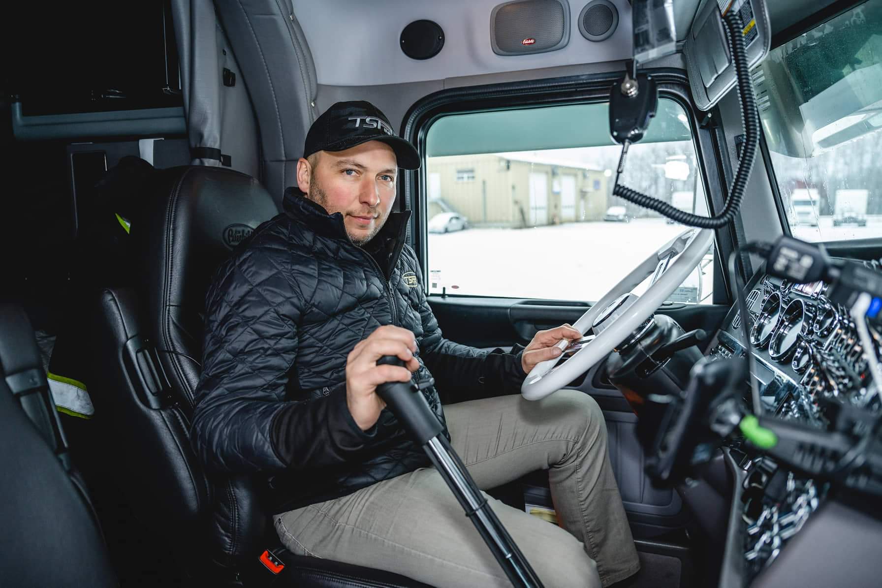 Un homme vêtu d'une veste noire et d'une casquette, assis sur le siège conducteur d'un camion, tenant le levier de vitesse et regardant la caméra.