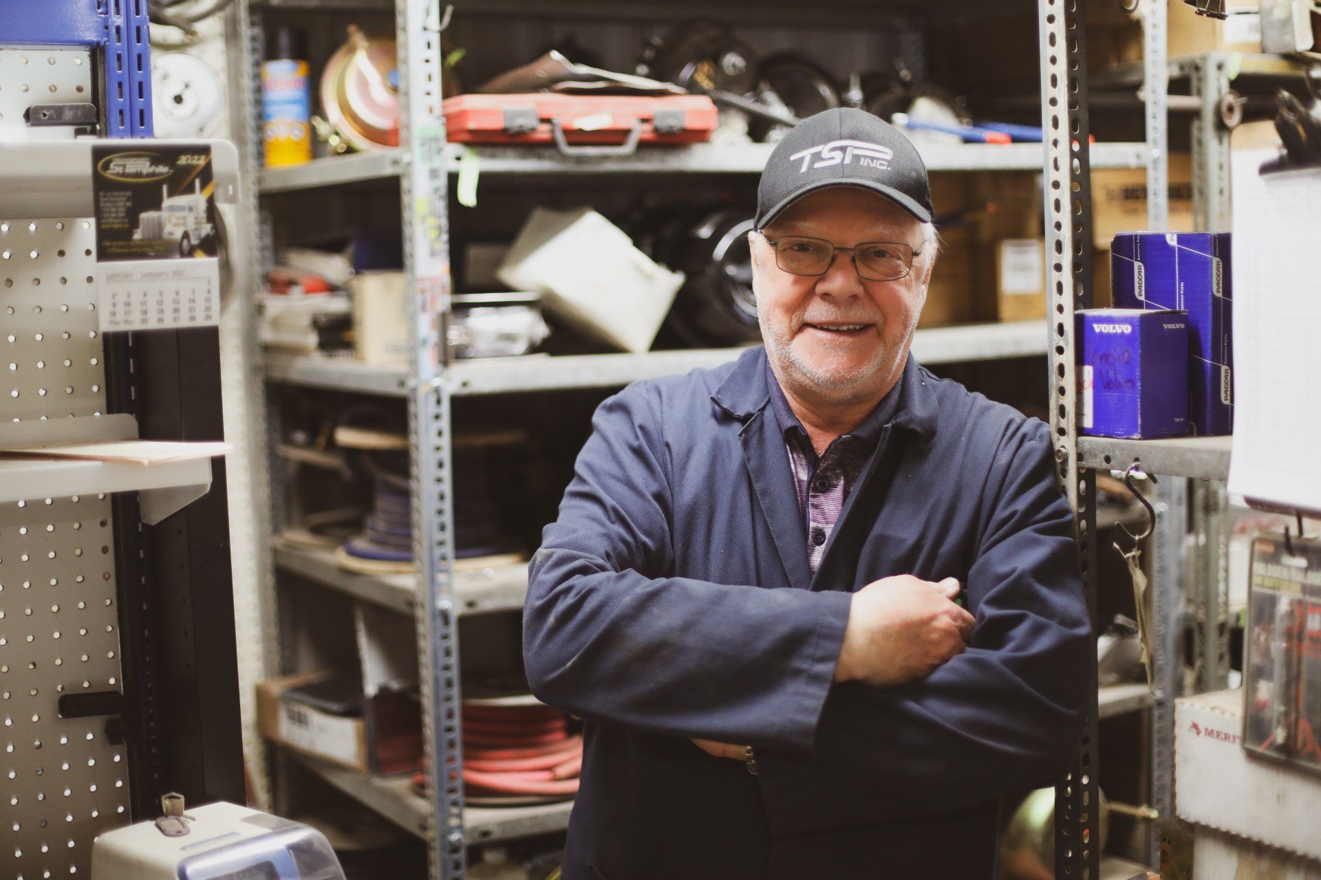 Une personne portant une veste sombre et une casquette de baseball se tient debout, les bras croisés, dans un atelier industriel avec des étagères de stockage.
