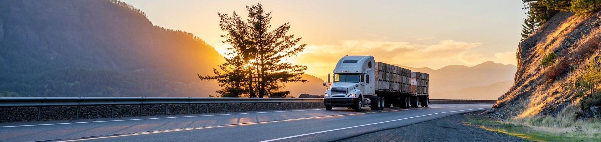 Semi-camion roulant sur une autoroute au coucher du soleil, montagnes et arbres en arrière-plan, lumière dorée.