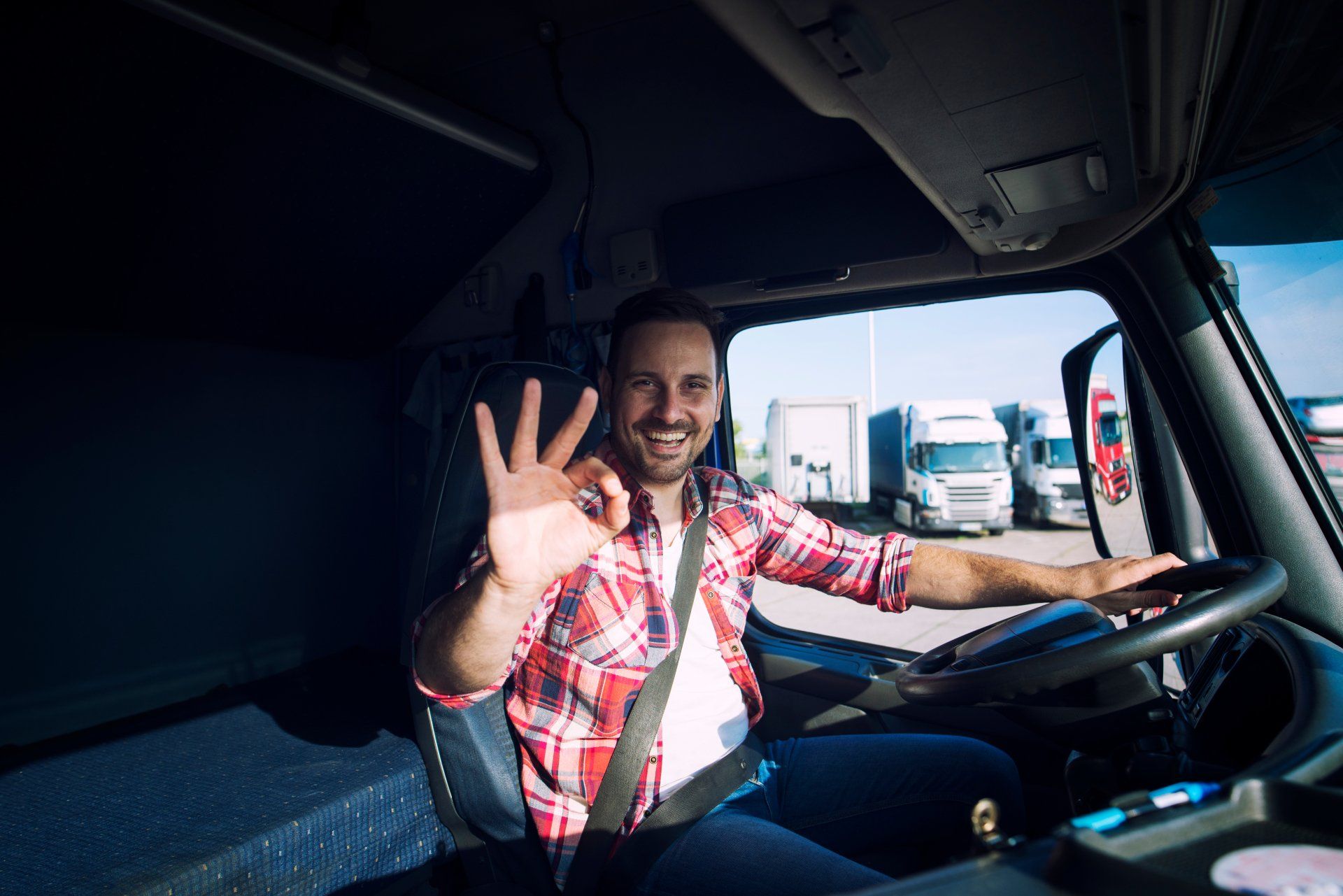 Un chauffeur de camion souriant, vêtu d'une chemise à carreaux rouges, est assis au volant et fait un geste de la main signifiant « OK ».
