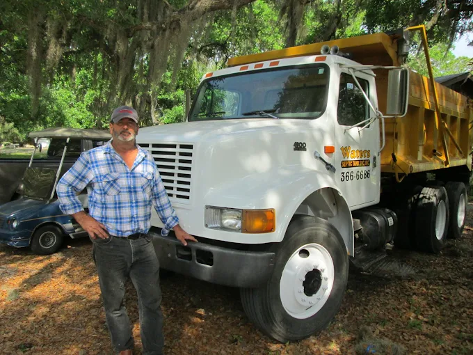 A man in a plaid shirt is standing in front of a dump truck.