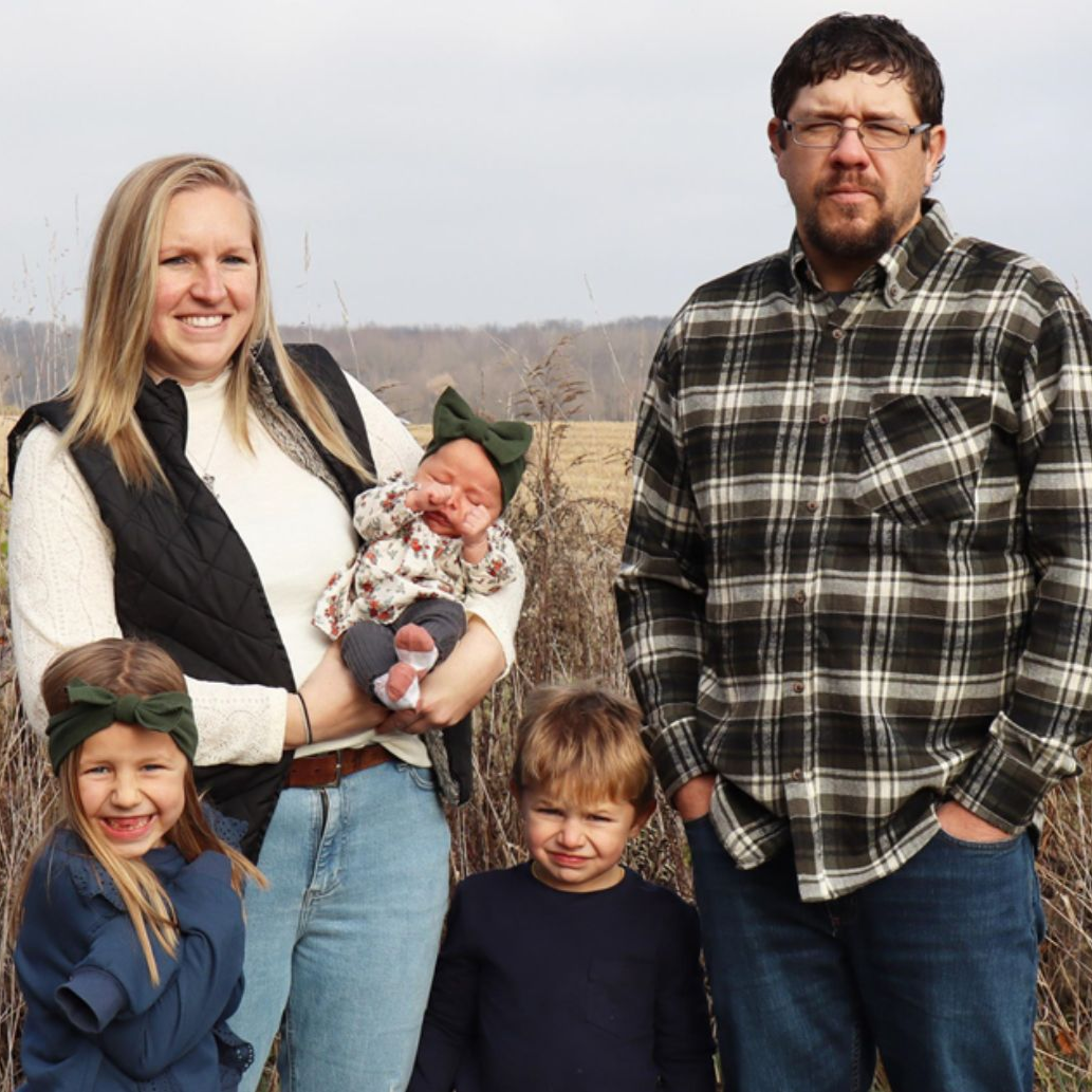 Family of five smiling outdoors. Mother holds baby, two children stand nearby, father smiles.