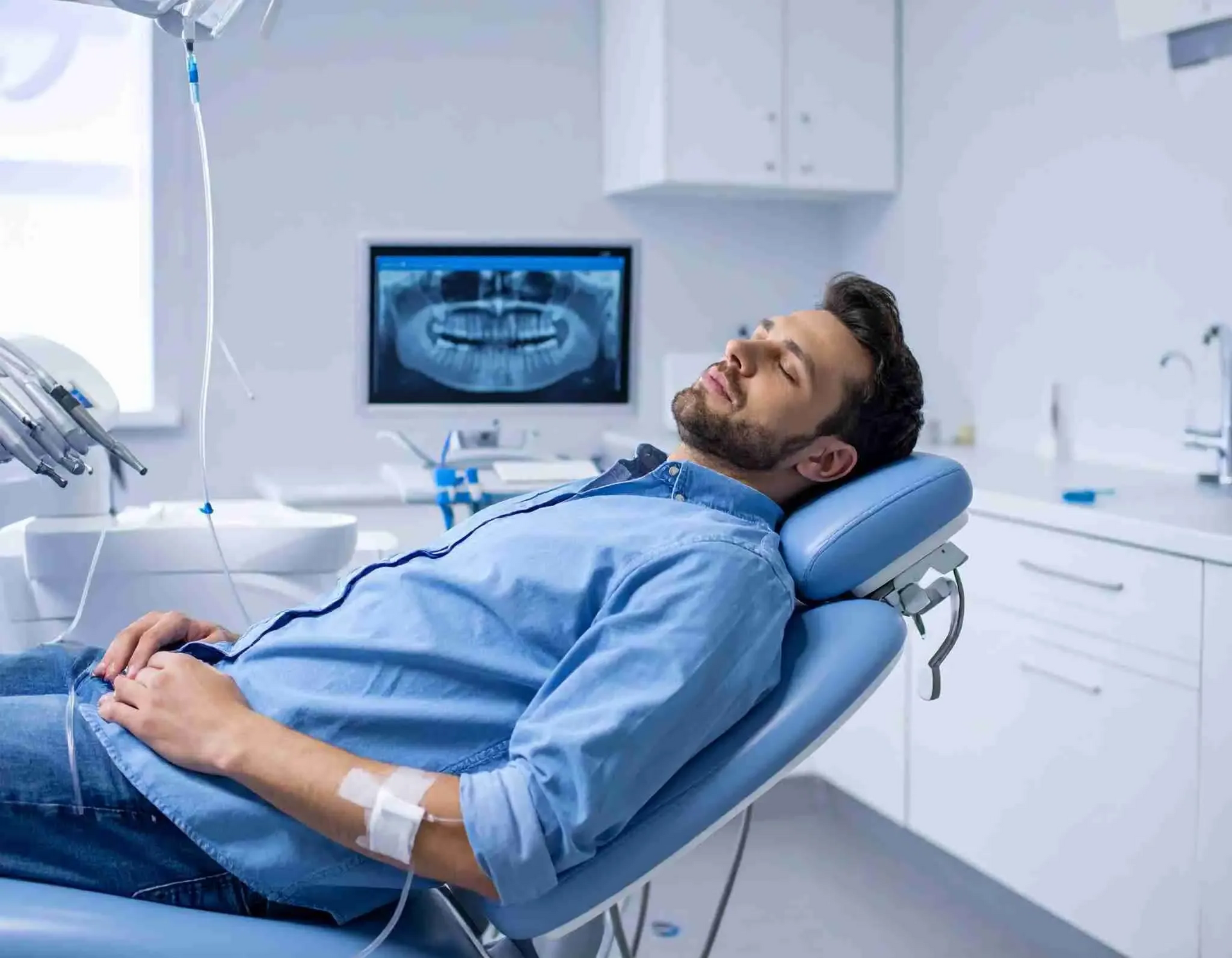 A person with an IV in their arm rests in a dental chair with a dental panoramic X-ray displayed on a monitor.