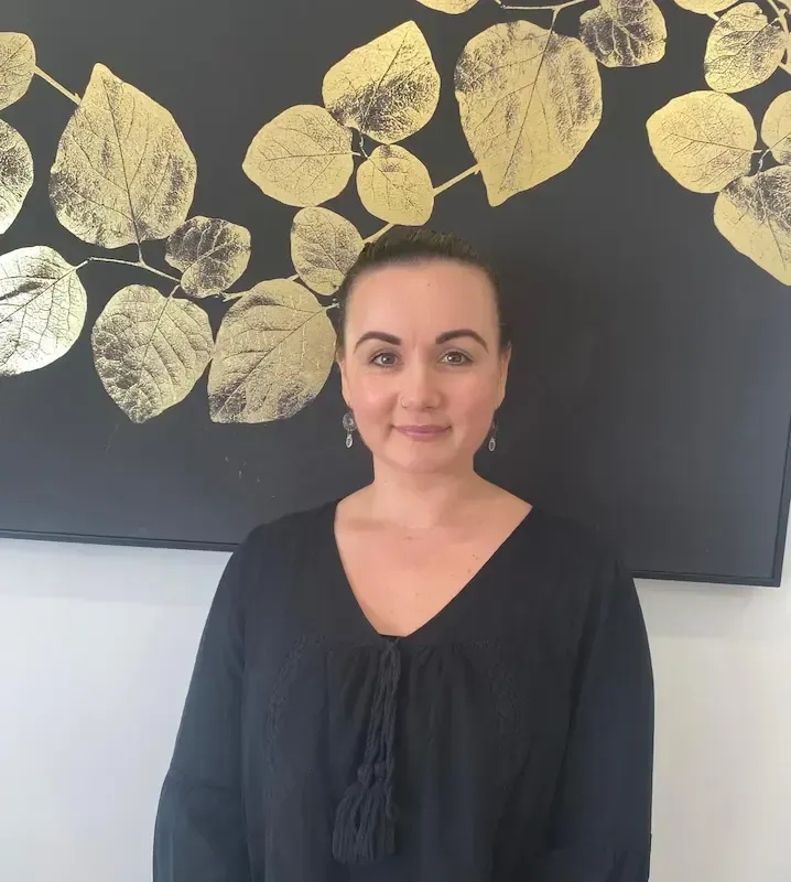 A person with long dark hair wearing a purple V-neck scrub top, standing against a white background.