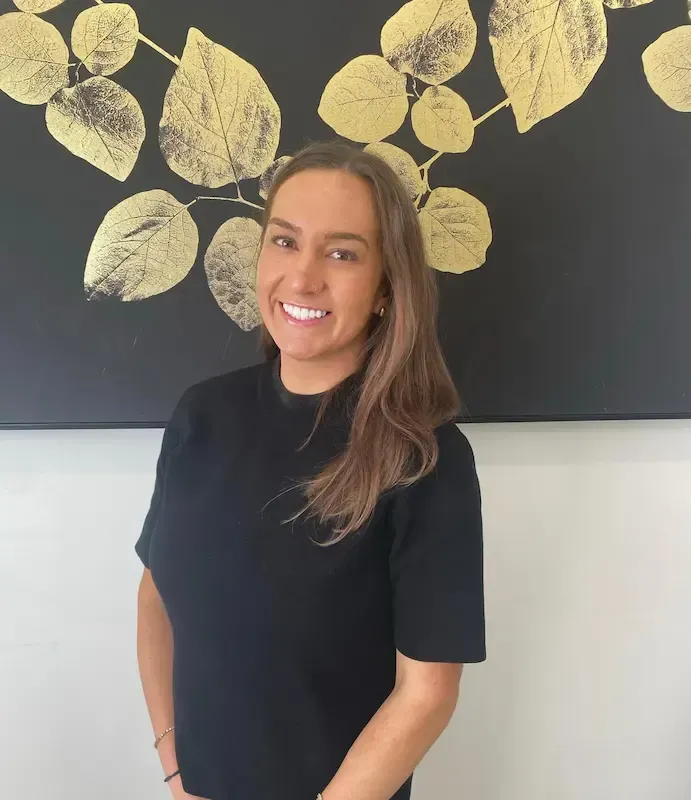 A person with long blonde hair and a black top, standing in front of a black wall decorated with gold leaf patterns.