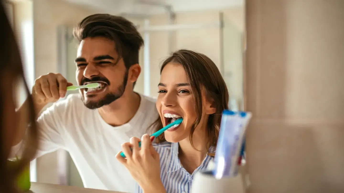 A couple brushes their teeth together in a bathroom, smiling at their reflections. The man is wearing a white shirt and has a beard; the woman is wearing a striped shirt.