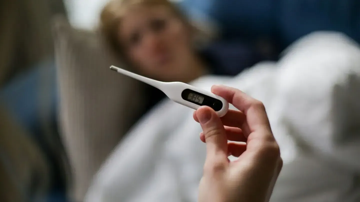 Hand holding digital thermometer, patient in bed with blurred background.