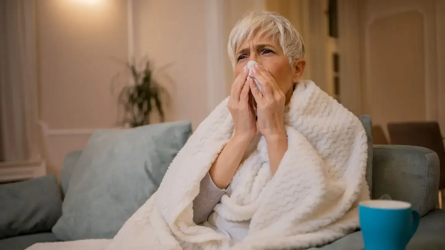 Woman with a cold blowing her nose, wrapped in a blanket, sitting on a couch with a mug nearby.