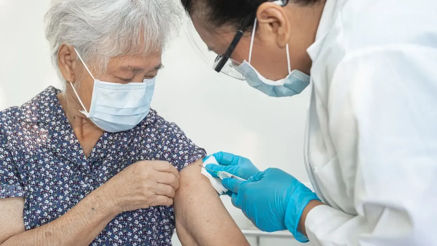 An elderly woman receives a vaccine in her arm from a healthcare worker in a medical setting.