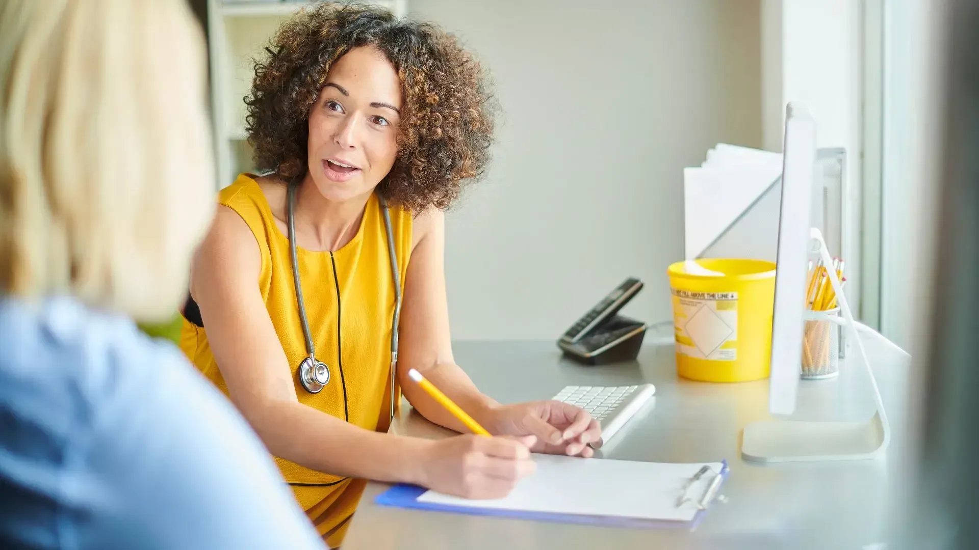 A doctor with curly hair in a yellow dress consults with a patient at a desk; stethoscope, sharps container present.