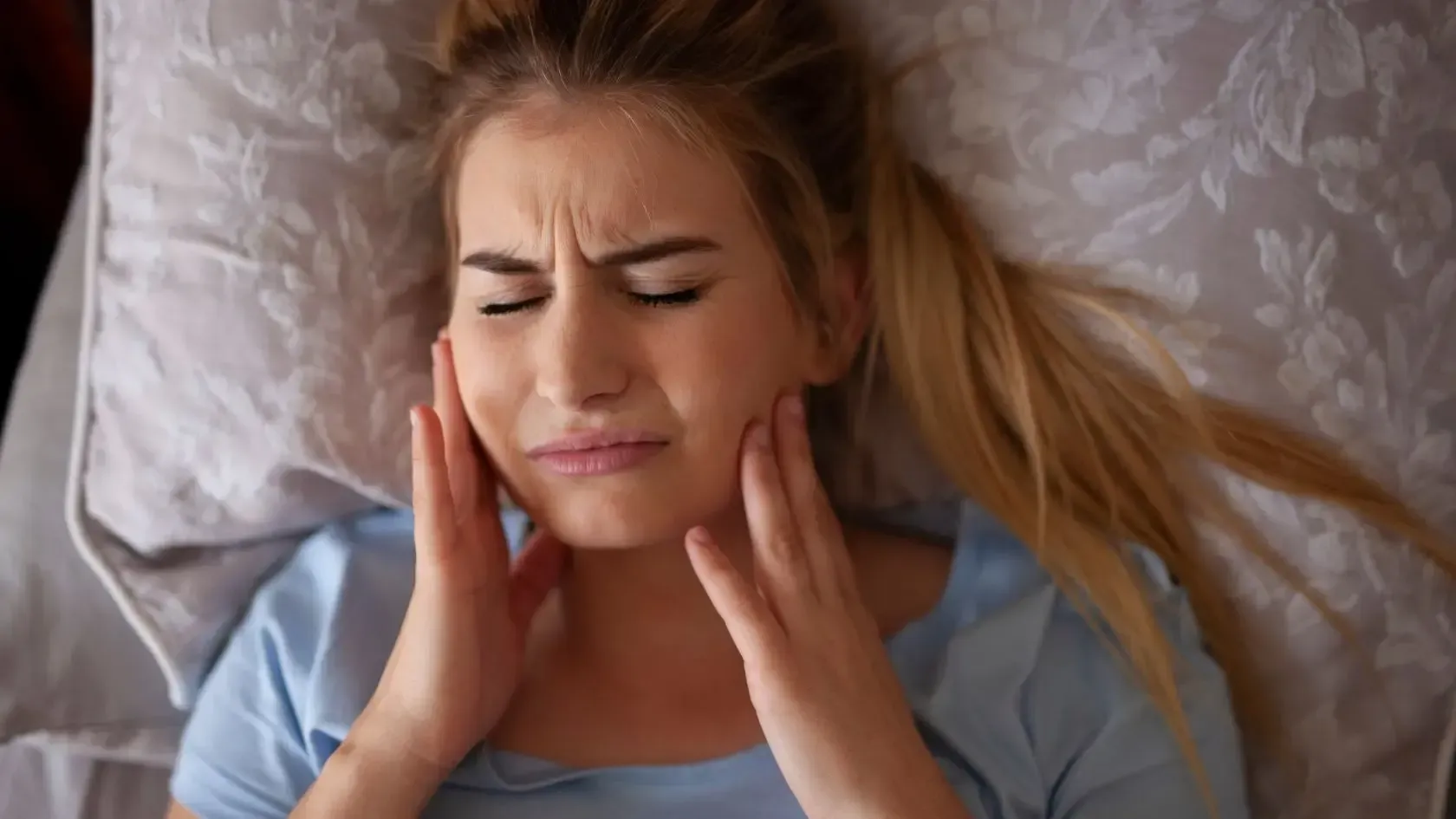 Woman in bed, grimacing, holding cheeks, possibly in pain.