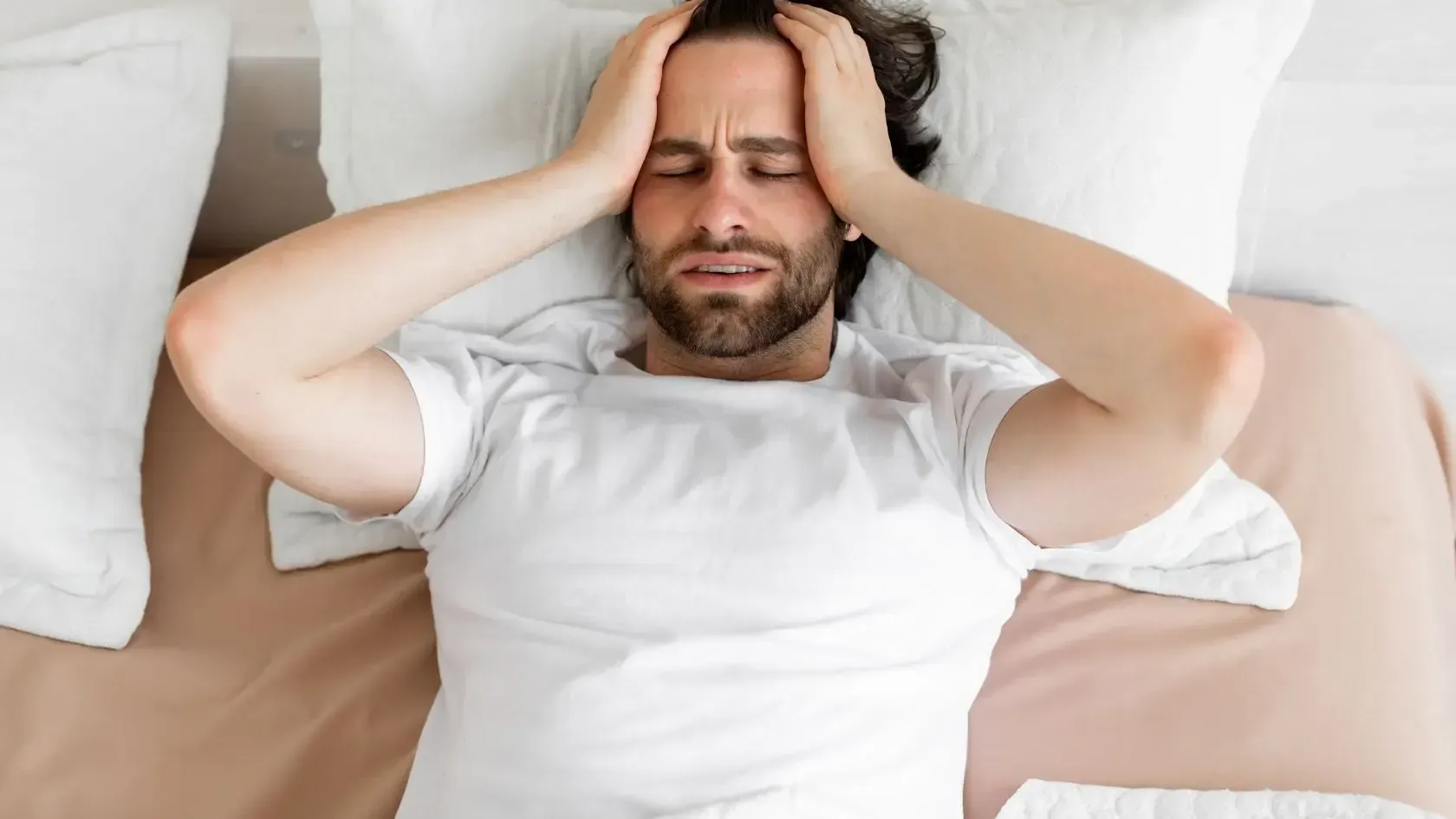 Man in bed with hands on head, eyes closed, looking distressed.