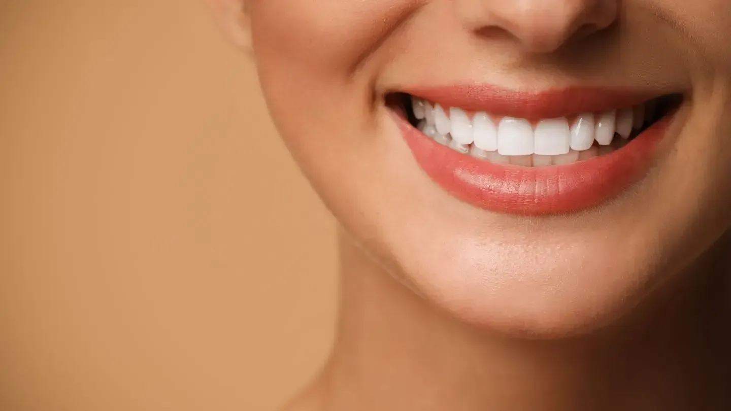 Close-up of a smiling mouth with bright white teeth and full, red lips, against a beige background.