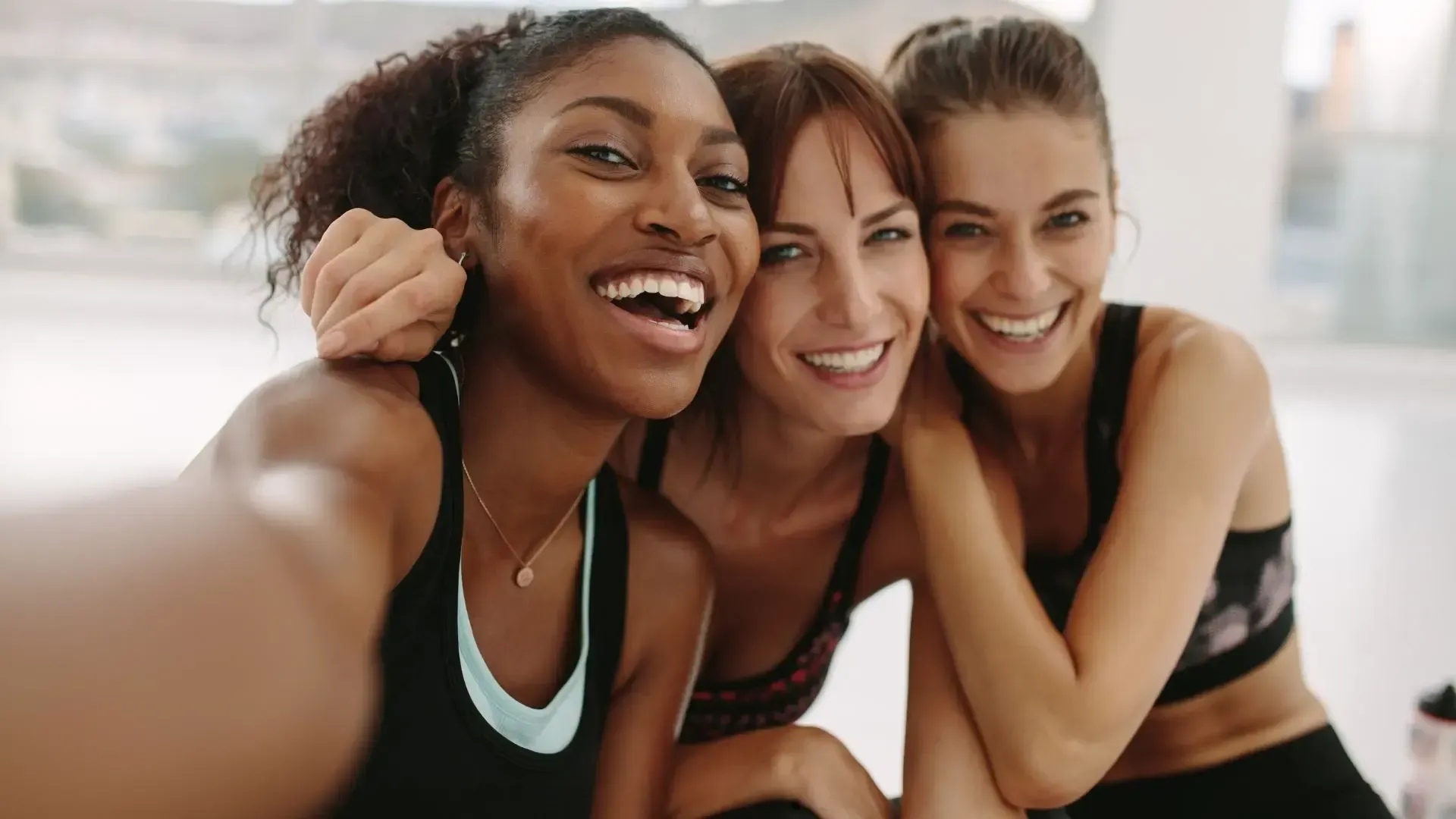 Three smiling women, embracing in a gym, taking a selfie.