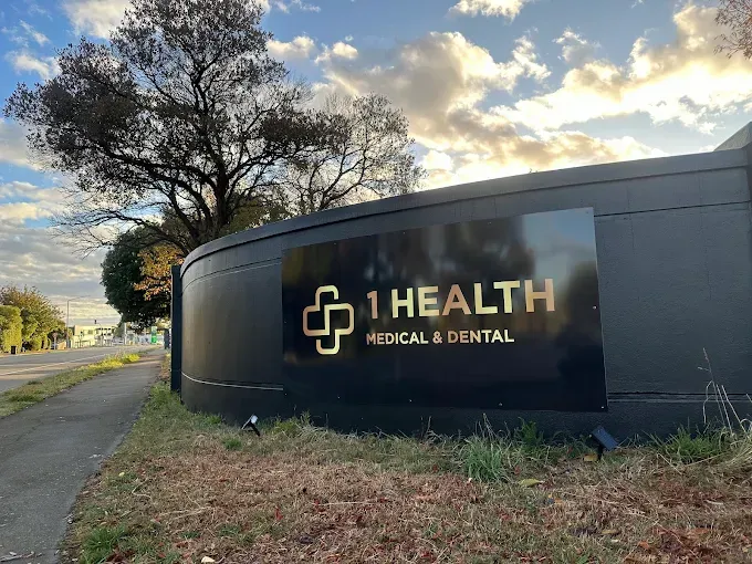Black sign for 1 HEALTH MEDICAL & DENTAL with gold lettering. Outdoor view, sign on curved wall, cloudy sky.