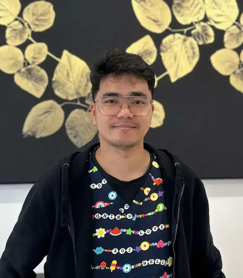 A person with glasses, a black hoodie, and a colorful patterned shirt poses in front of a black wall with gold leaf art.