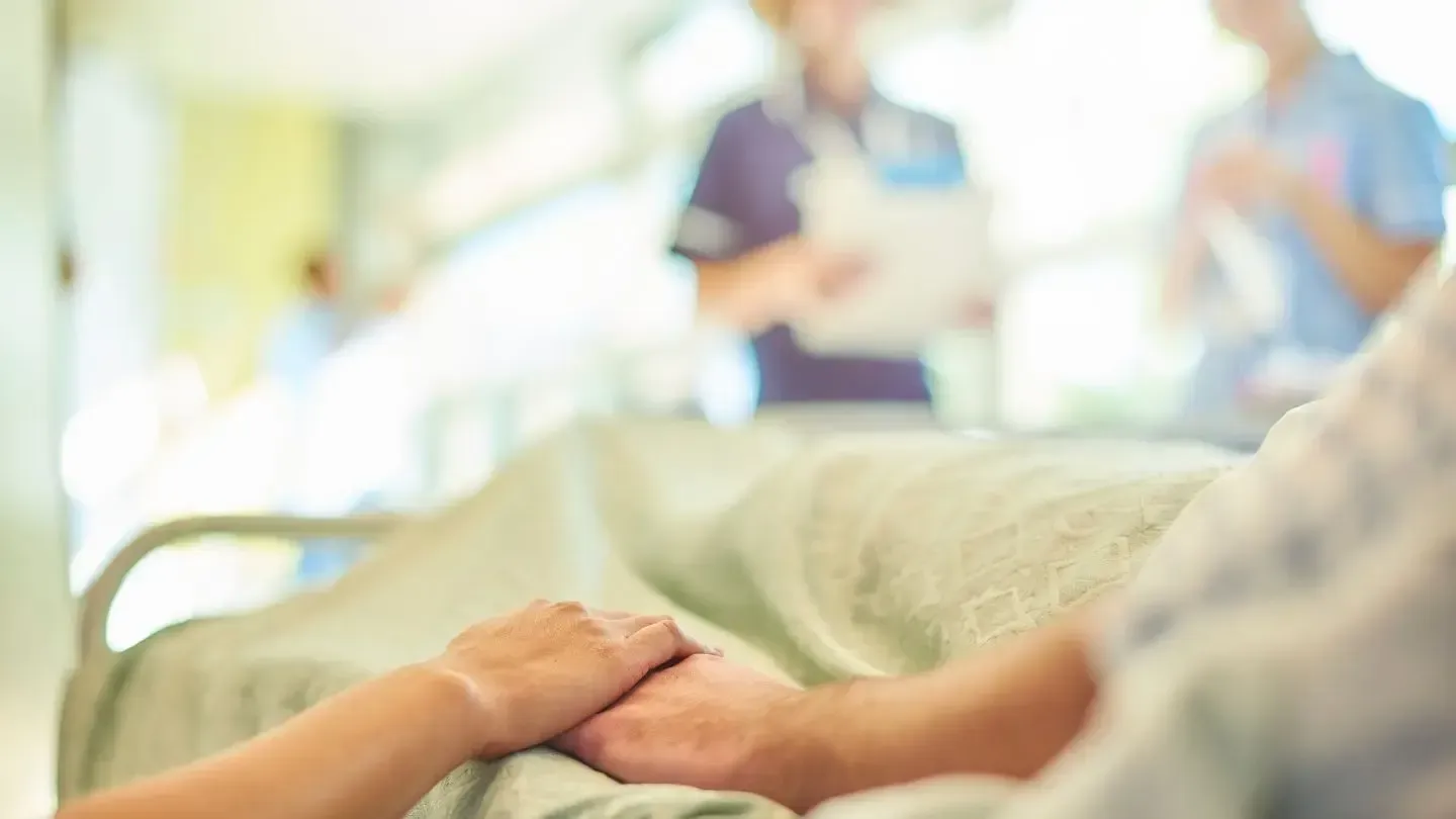 Person holding the hand of another in a hospital bed, with blurred medical staff in the background.