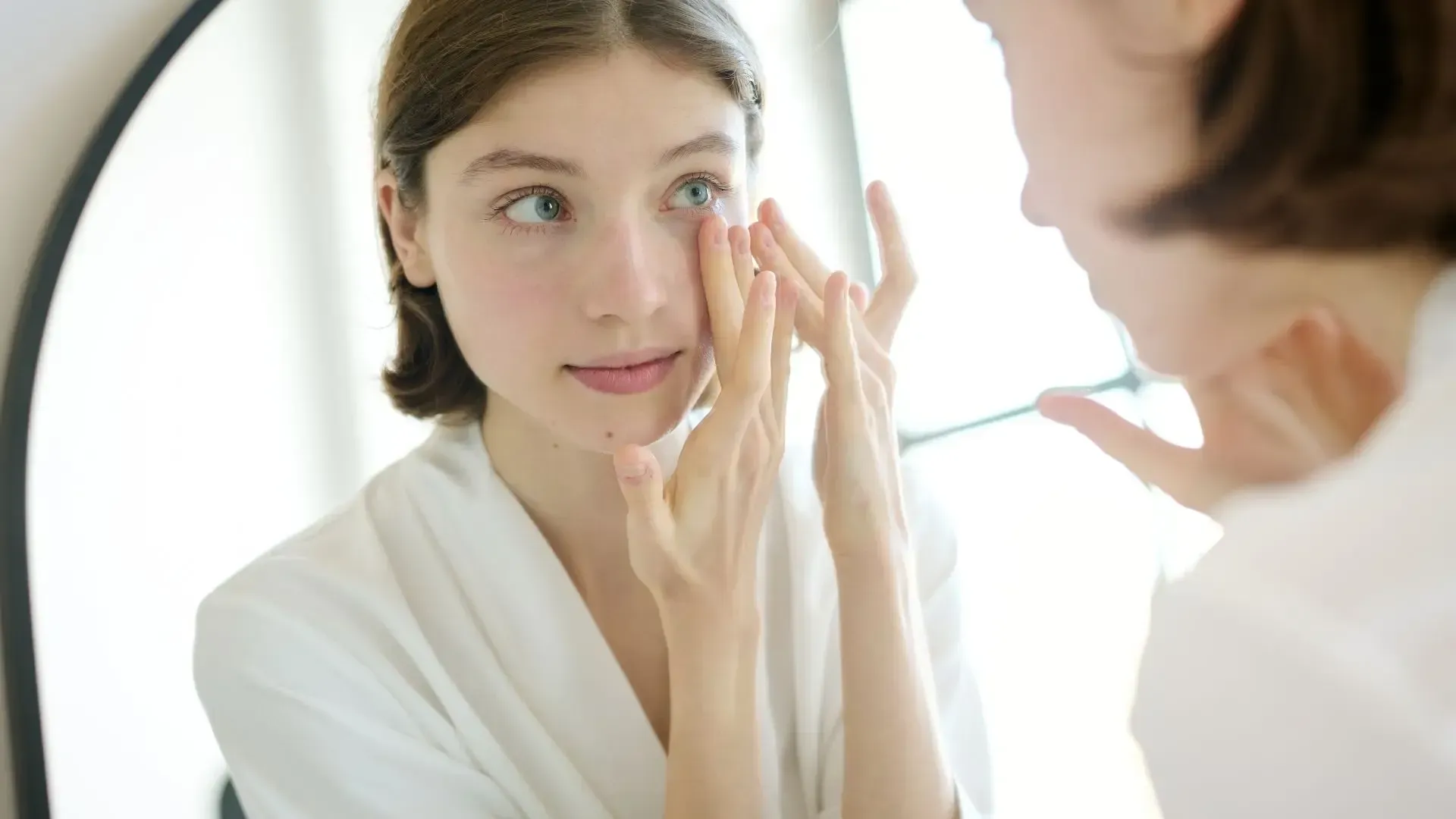 Woman in white robe looking in mirror, touching face with fingers.