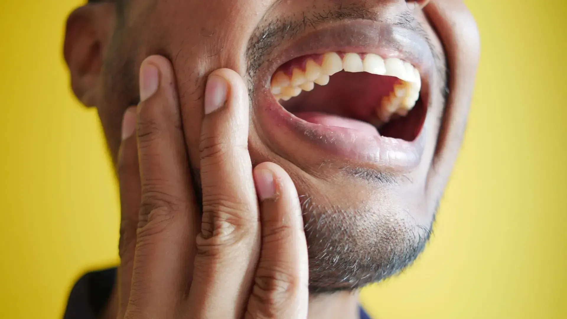 Man with hand on cheek, mouth open wide, possibly in pain, against yellow background.