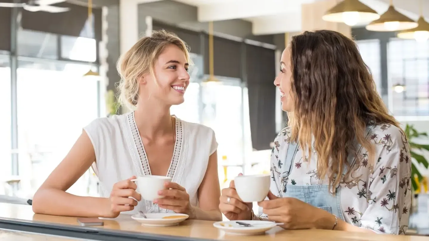 Two women smiling, sitting at a cafe table, holding coffee cups. Natural light illuminates the setting.