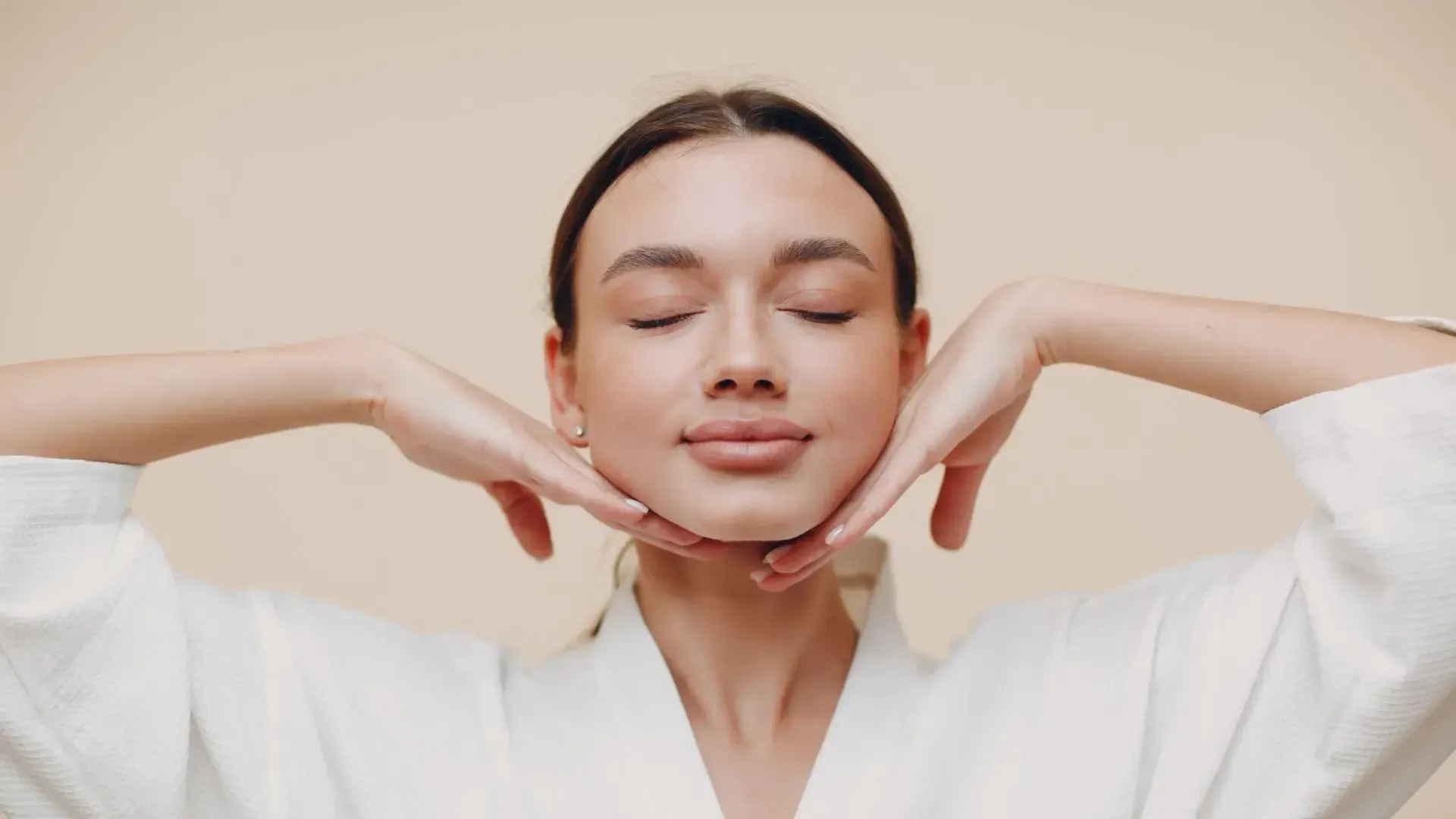 Woman with closed eyes, hands framing face. Wearing white robe, neutral background.