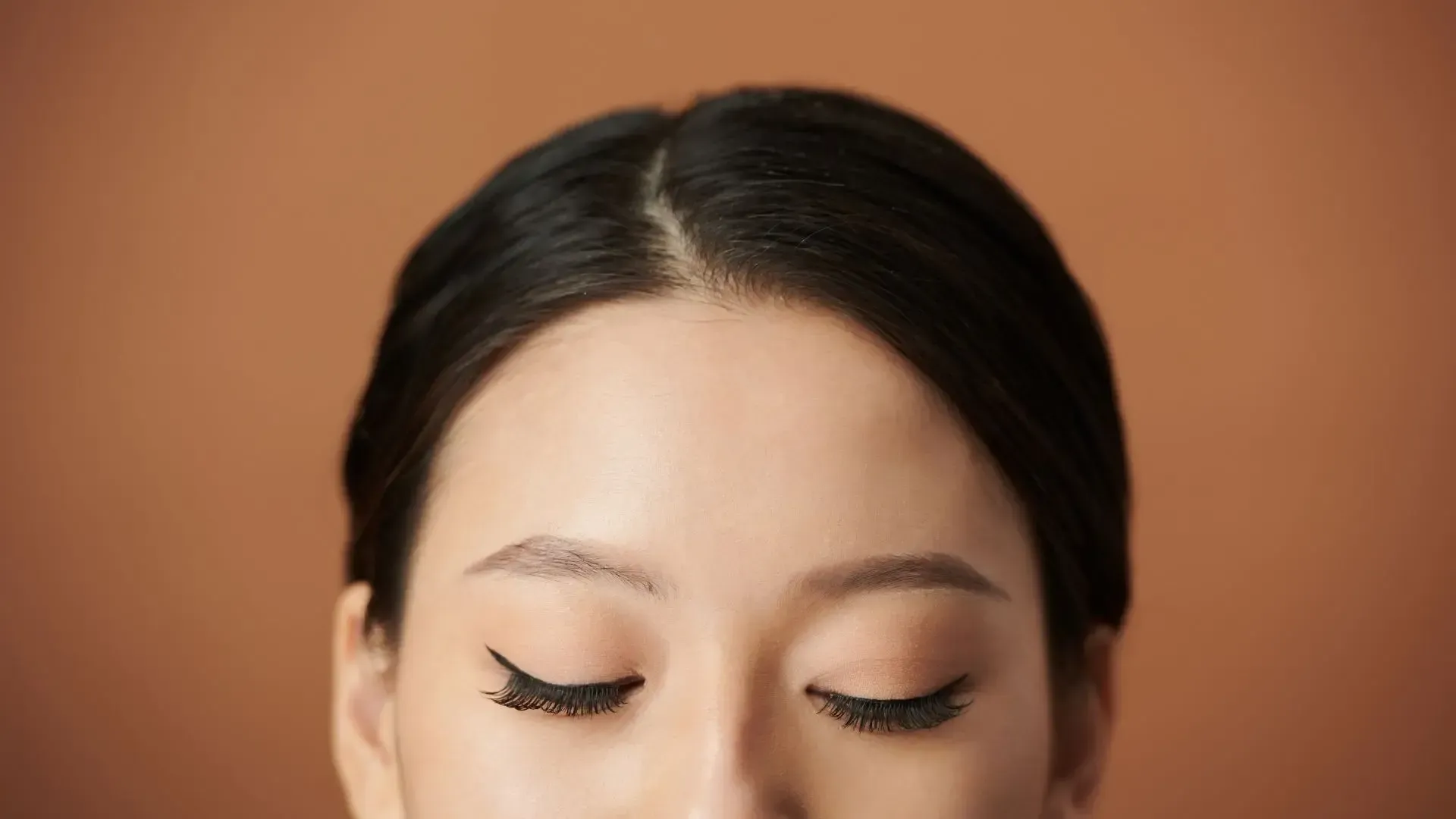 Woman with dark hair parted down the middle, eyes closed. Beige background.