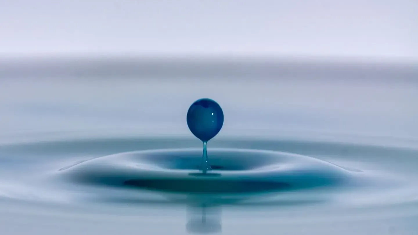 Blue droplet suspended above a ripple in water.