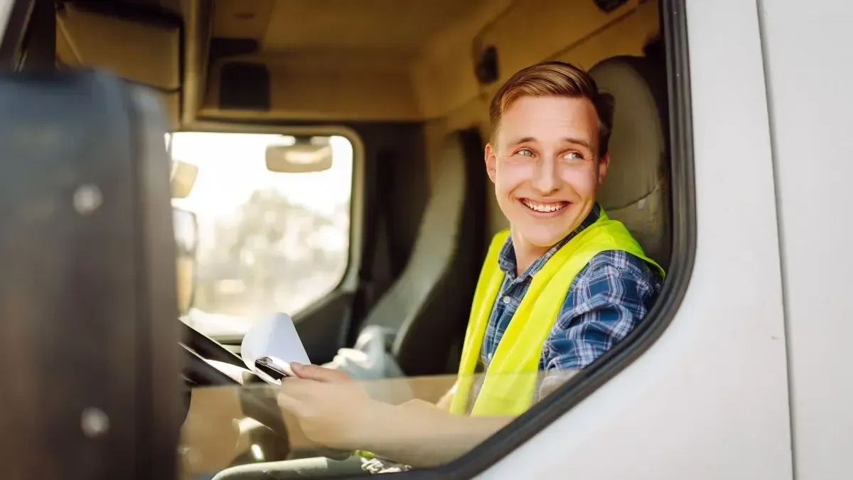 Man in truck cab wearing a safety vest, smiling and looking to the side.
