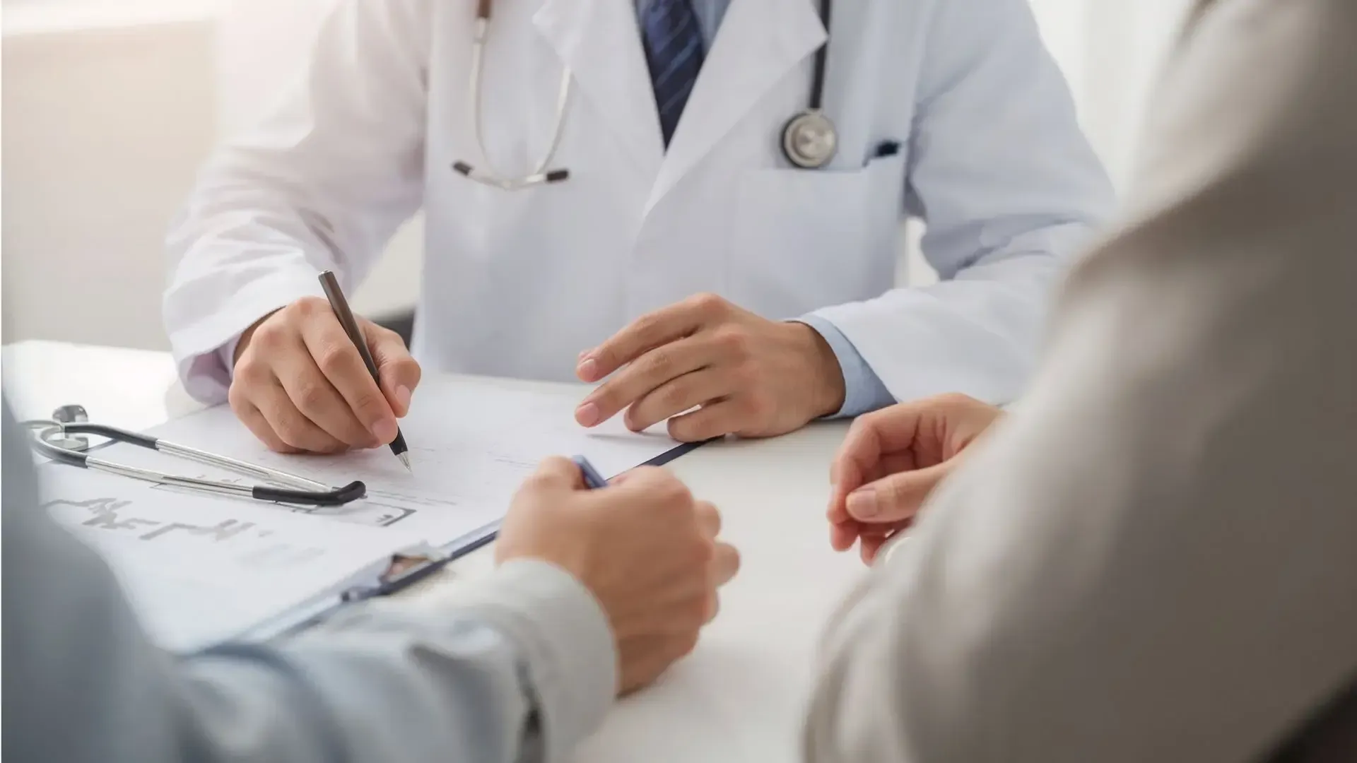 Doctor writing on paperwork with a patient; stethoscope on table.