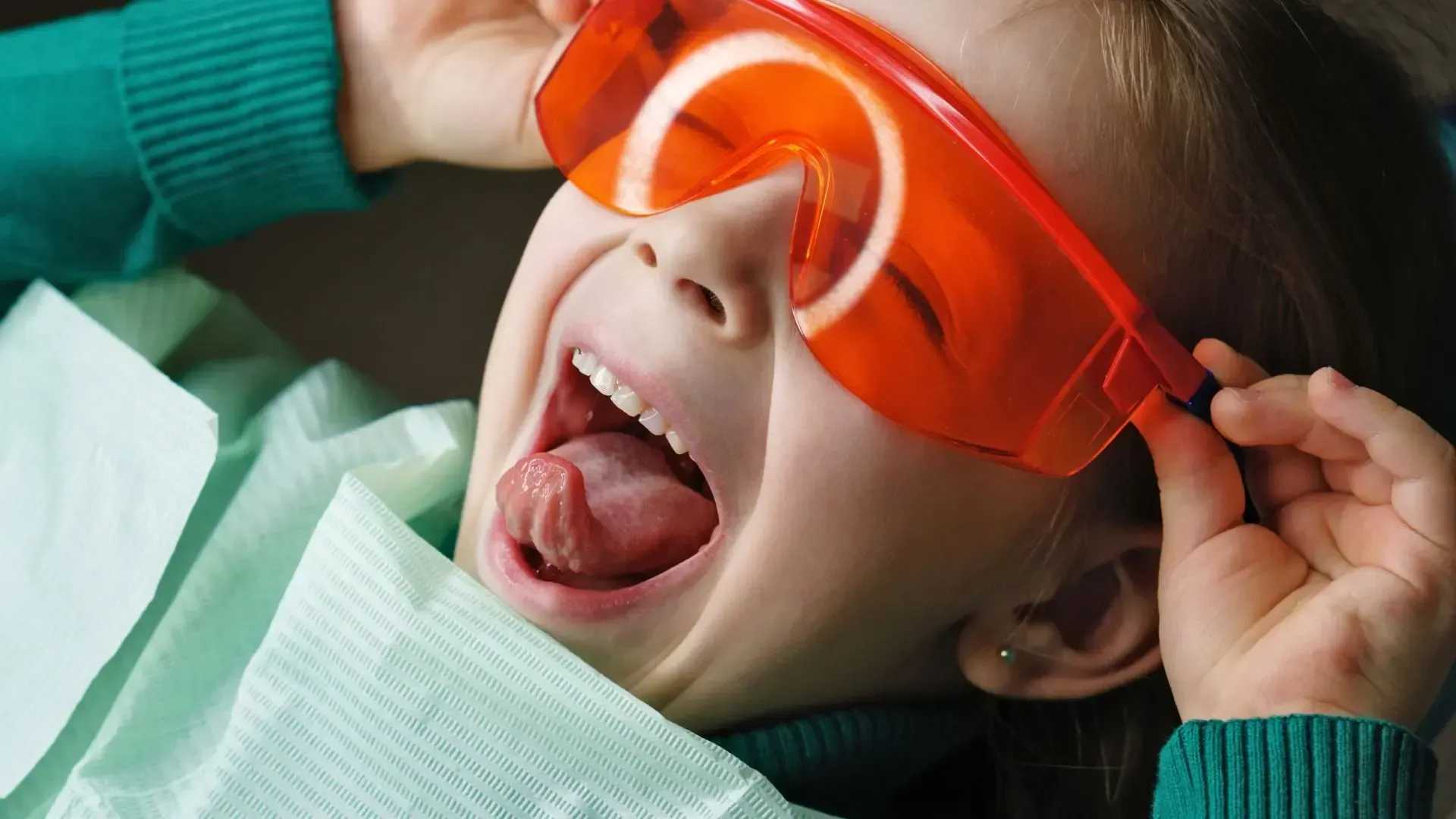 A person in a dental chair wearing orange safety goggles has their mouth wide open.