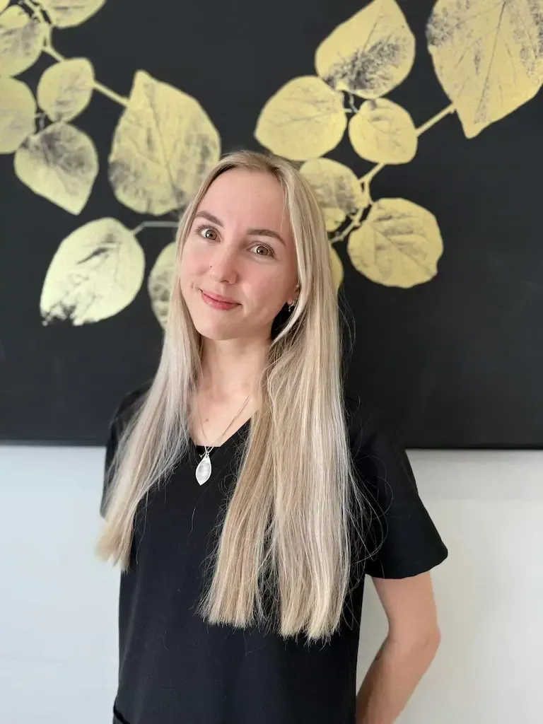 A person with long blonde hair and a black top, standing in front of a black wall decorated with gold leaf patterns.