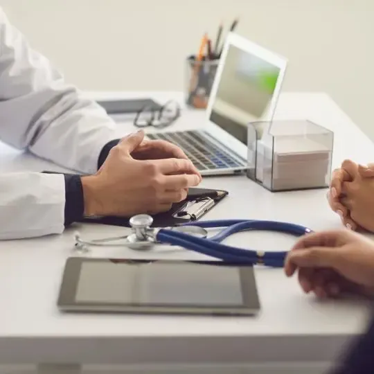 A doctor is talking to a patient at a desk with a laptop and a tablet.