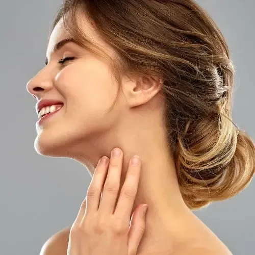 Woman smiling, touching her neck; brown hair in a bun, against a gray background.