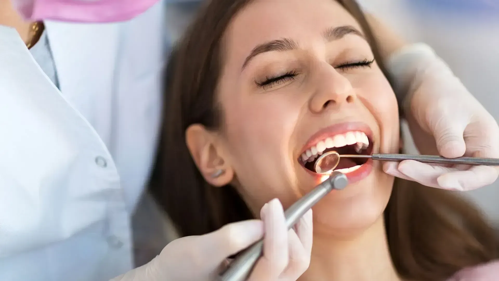 A patient with eyes closed receives a dental examination by a professional using a dental mirror and handpiece.