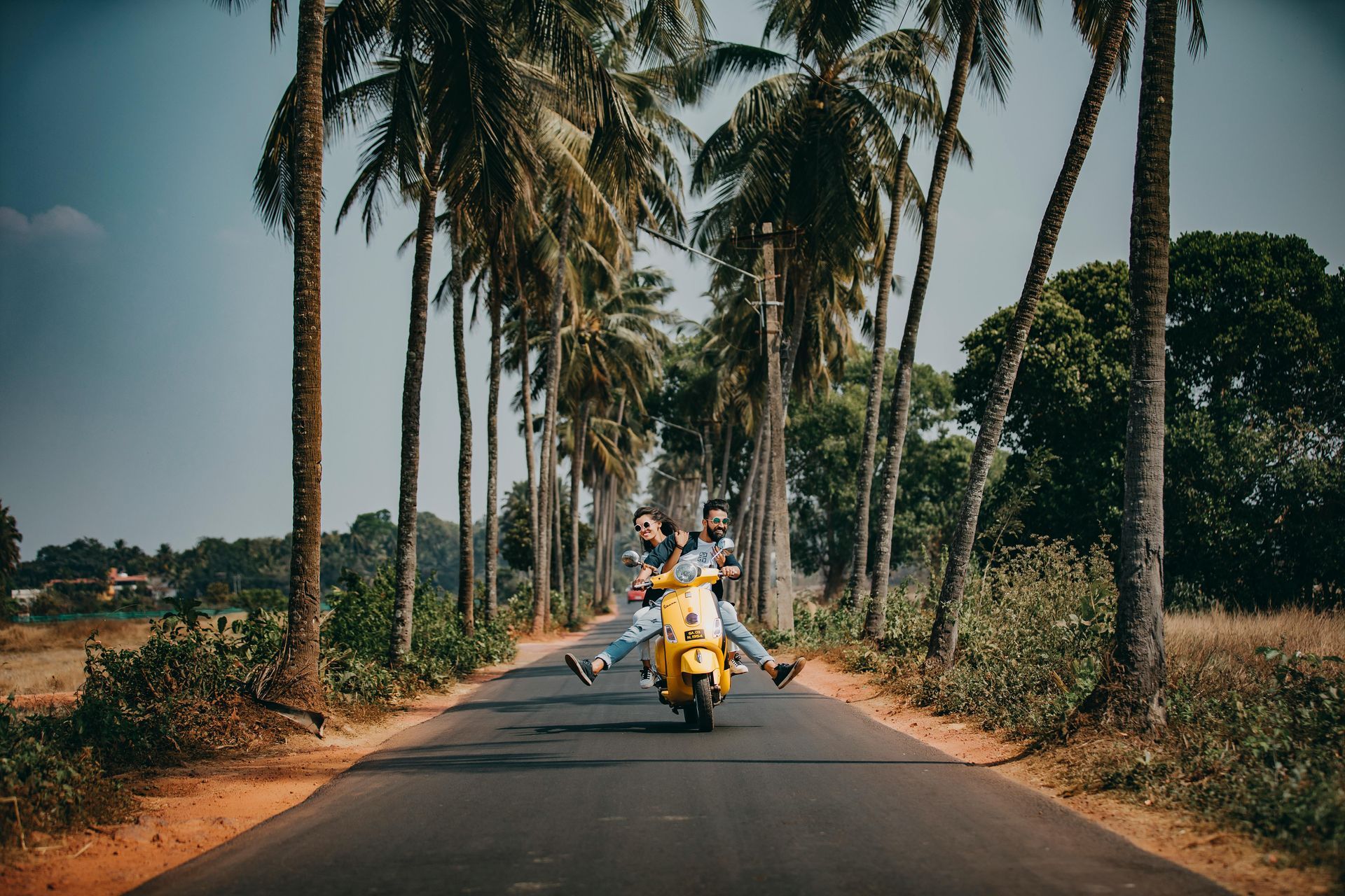 Two people joyfully ride a yellow scooter down a road lined with palm trees.