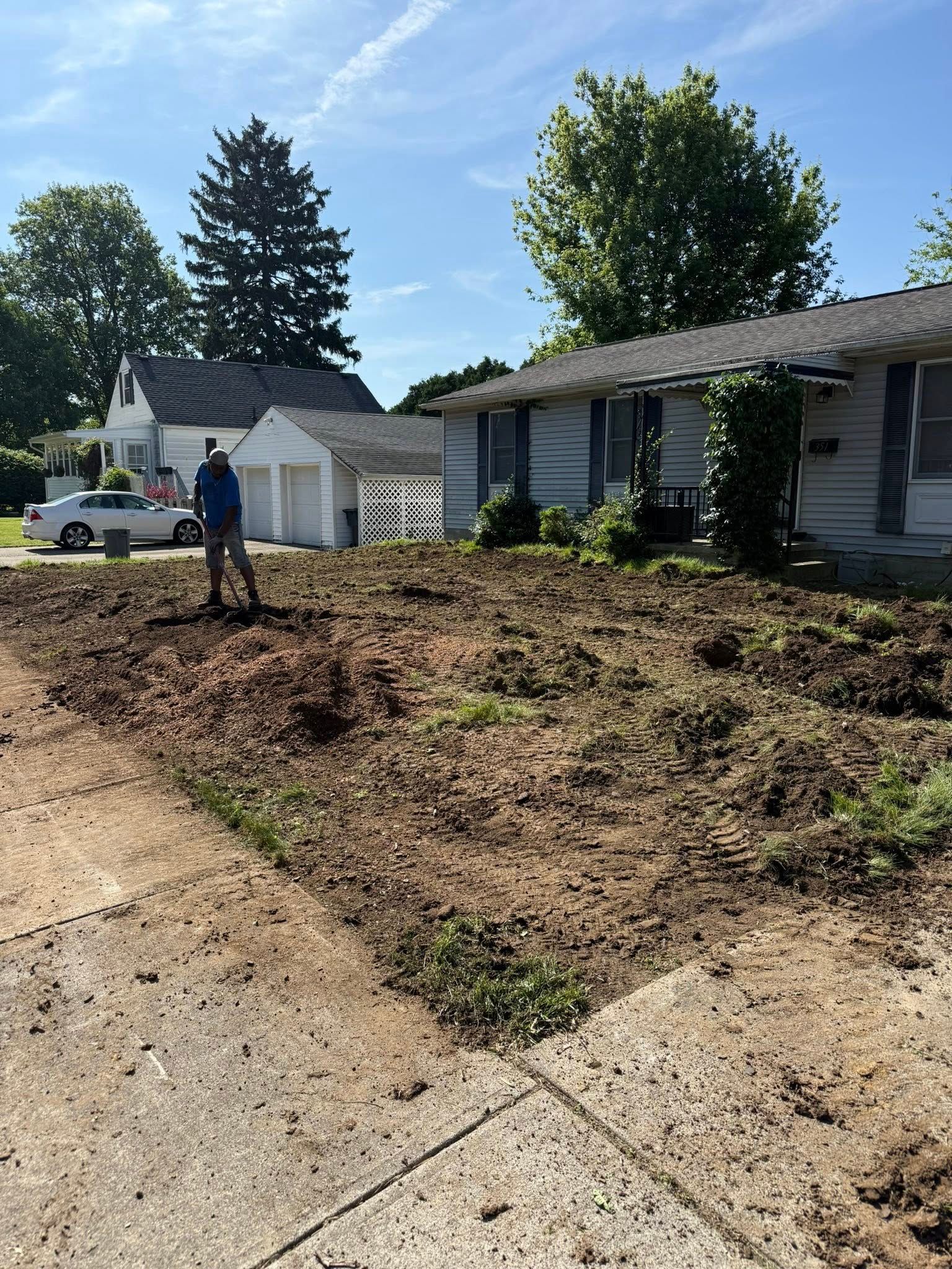 A person standing in a dirt yard in front of a house, preparing a garden. Sunny day.